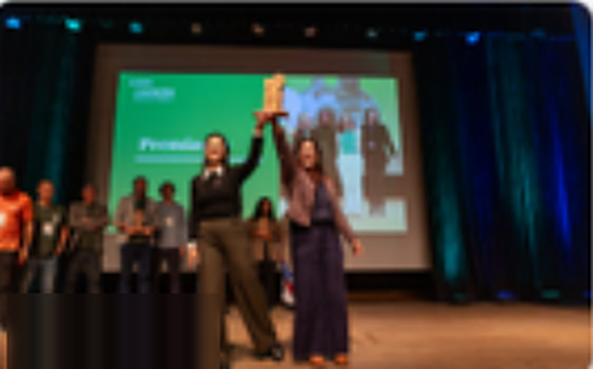 Duas pessoas no palco erguem um troféu diante de um telão, com os demais finalistas ao fundo.